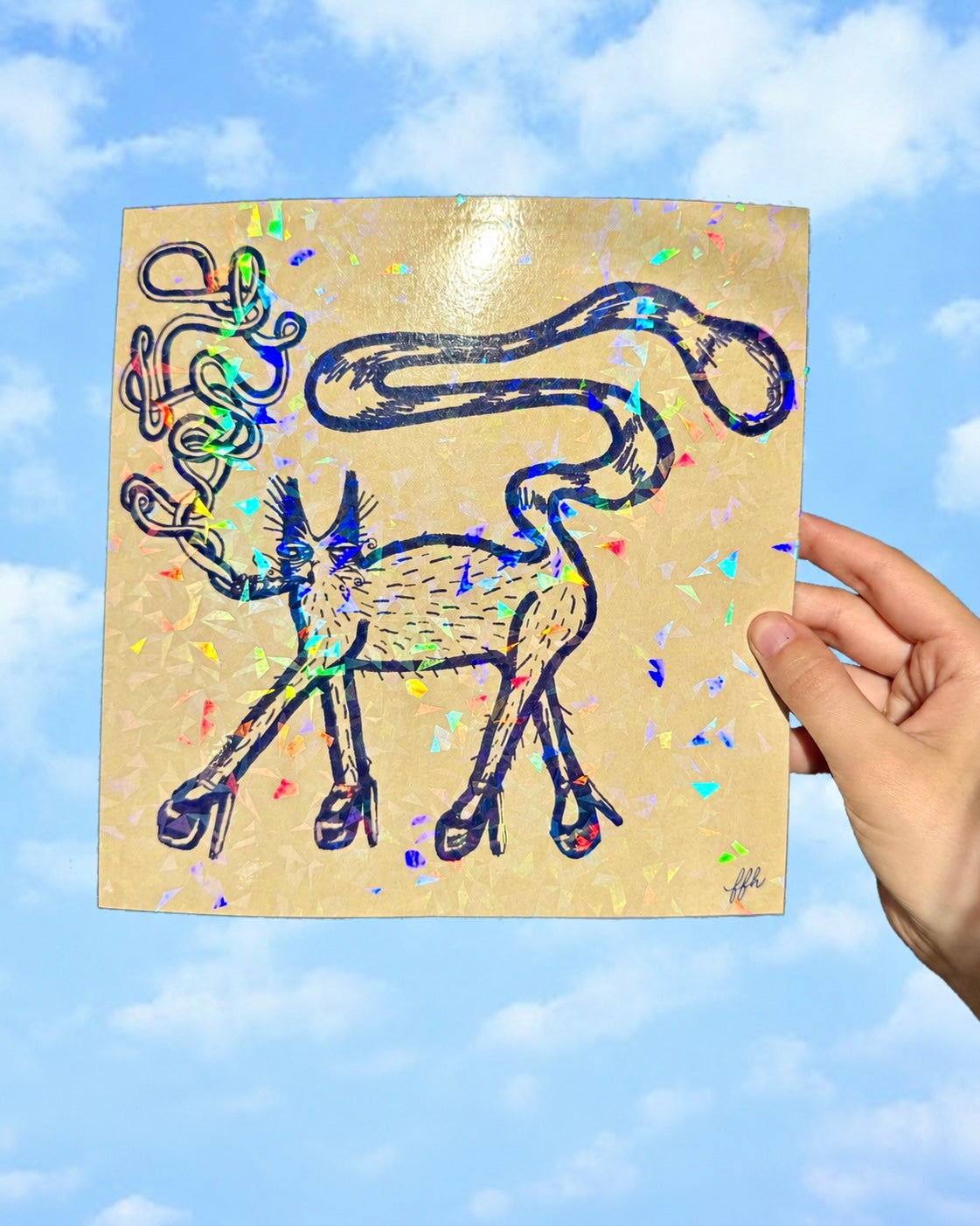 A hand holds a square gold-toned art print or card featuring a whimsical black line drawing of a cat with abstract, looped limbs and tail, overlaid with iridescent confetti, against a blue sky background with clouds.