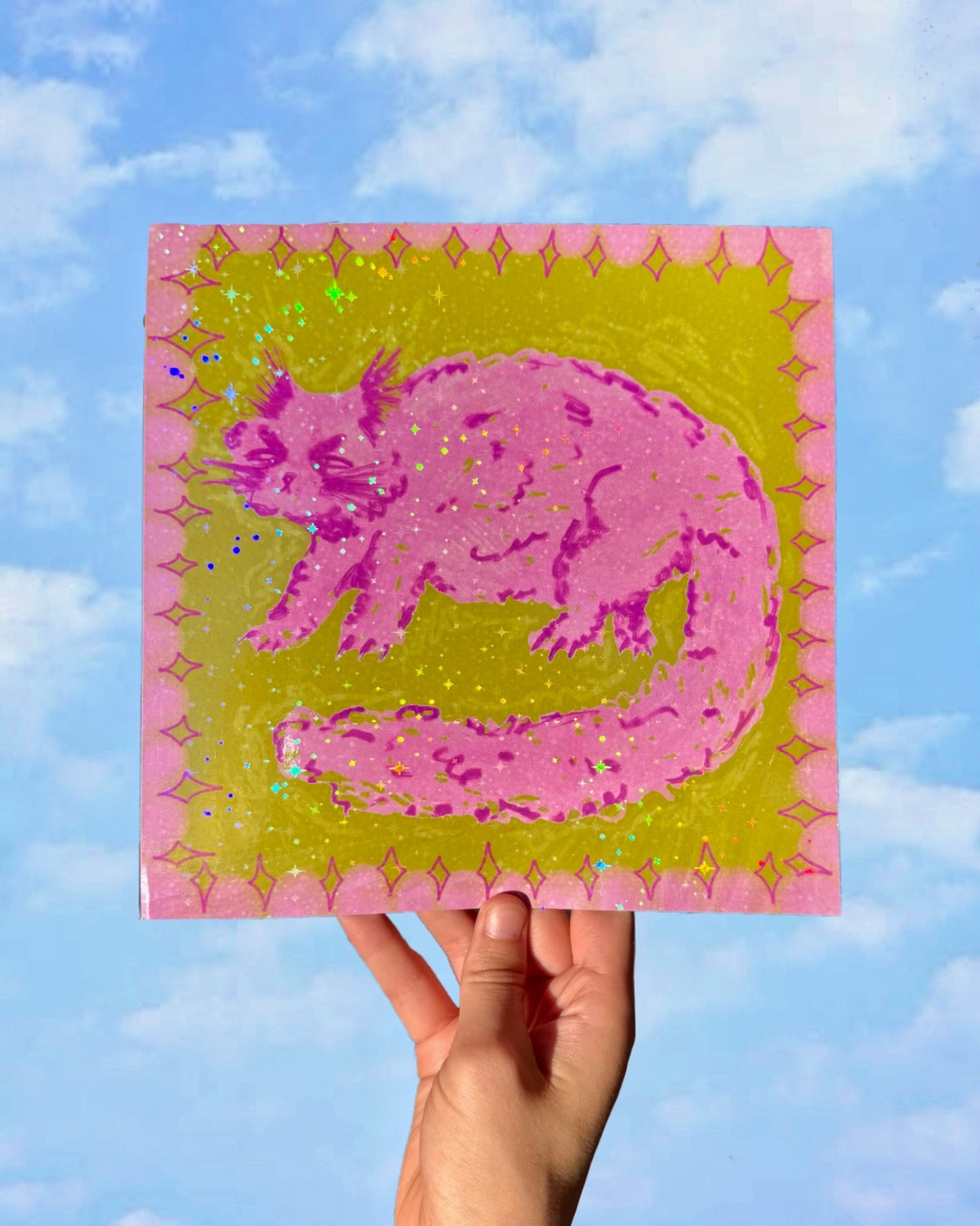 A hand holds a square glass art piece featuring a pink cat silhouette on a yellow-green background with a pink patterned border, set against a blue sky with clouds.